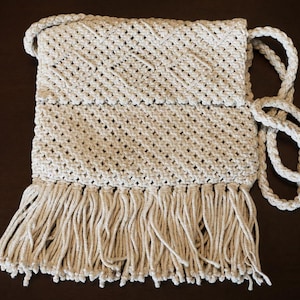 May include: A cream-colored macrame crossbody bag featuring a diamond-patterned knot design, a fold-over flap, and a long twisted rope shoulder strap. The bottom edge of the bag is finished with a thick row of long, hanging fringe tassels.
