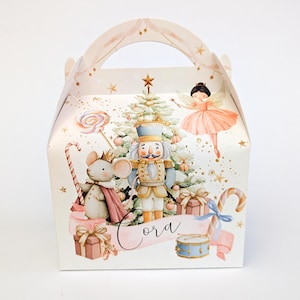 May include: A decorative gift box with a handle, featuring a festive Nutcracker theme. The box showcases a Christmas tree, a fairy, a mouse, and a nutcracker soldier. The name "Cora" is written on a banner.
