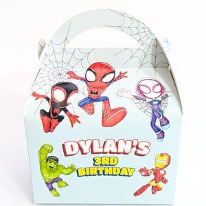 May include: A light blue cardboard favour box with a handle, featuring cartoon illustrations of Spider-Man, Ghost-Spider, Miles Morales, Hulk, and Iron Man. The text "DYLAN'S 3RD BIRTHDAY" is printed in red and yellow on the front of the box.