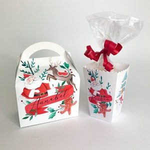 May include: Two white Christmas gift boxes. One box has a handle and features Santa Claus, a reindeer, and the name "Annabell" on a red banner. The other box has a clear cellophane bag tied with a red ribbon and features Santa Claus and the name "Reuben".