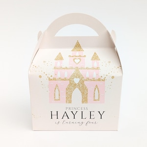 Peut inclure: Une boîte à cadeaux rose clair avec un château doré scintillant. La boîte a une poignée et le texte "Princess Hayley is turning four".