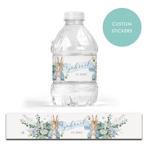 May include: A clear plastic water bottle with a custom label featuring a watercolour illustration of a rabbit in a blue jacket. The label reads "Gabriel IS ONE" with floral accents. A matching label is below.