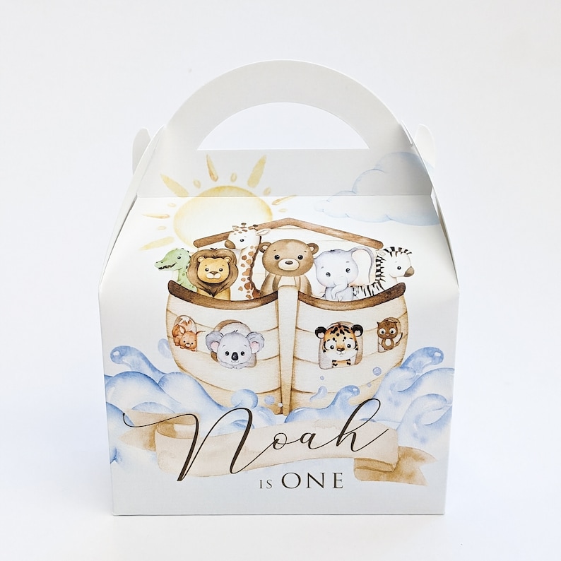 May include: White party favour box with a watercolour illustration of Noah's Ark, featuring various animals. The box has a handle and includes the text "Noah is One" in a script font, ideal for a first birthday celebration.