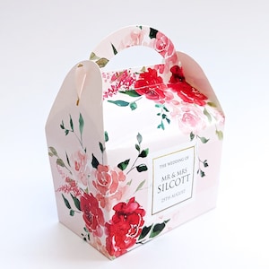 May include: A pink and white floral patterned favour box with a handle. The box has a white label with the text "The Wedding of Mr. & Mrs. Silcott 25th August".