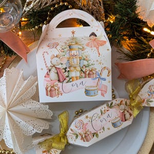 Può includere: Una scatola regalo bianca e un cracker decorati con il tema natalizio dello Schiaccianoci. La scatola e il cracker presentano illustrazioni di uno schiaccianoci, un topo, una ballerina e dei regali. Il nome "Cora" è scritto su uno striscione. Nastro dorato e centrino di carta.
