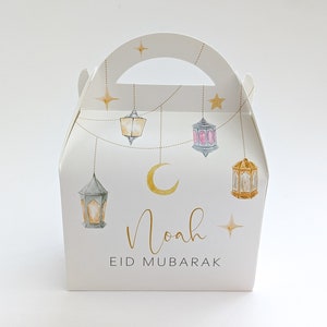 May include: White favor box with a handle, decorated with gold stars, a crescent moon, and hanging lanterns. The box has the name "Noah" and the text "Eid Mubarak" printed on it.