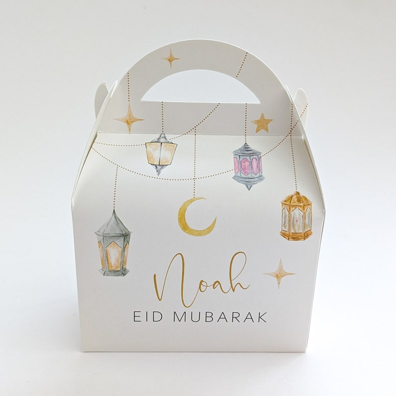 May include: White favor box with a handle, decorated with gold stars, a crescent moon, and hanging lanterns. The box has the name "Noah" and the text "Eid Mubarak" printed on it.