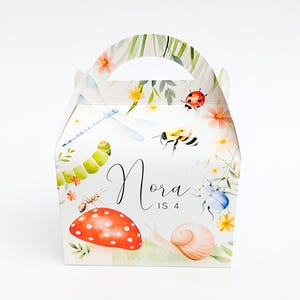 Puede incluir: Una caja de regalo blanca con asa, decorada con ilustraciones de acuarela de insectos, flores y una seta roja. El nombre "Nora" y el número "4" están escritos en la parte delantera.