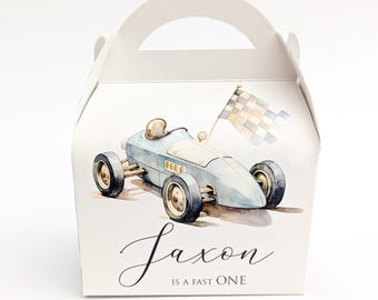 May include: A white favor box with a watercolor illustration of a vintage blue race car and a checkered flag. The text "Saxon IS A FAST ONE" is written in a script font below the car. The box has a handle.