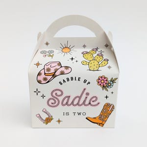 Op de afbeelding: Witte feestartikelen doos met een handvat, versierd met een roze cowboyhoed, cactus, bloemen en een cowboylaars. De tekst "Saddle Up Sadie is Two" wordt weergegeven in roze en zwart.