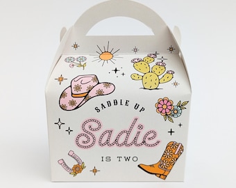 Puede incluir: Caja de regalo blanca con asa, decorada con un sombrero de vaquero rosa, cactus, flores y una bota de vaquero. El texto "Saddle Up Sadie is Two" se muestra en rosa y negro.