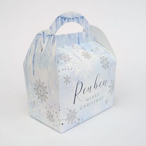 Könnte beinhalten: Hellblaue Geschenkbox mit Griff, verziert mit silbernen Schneeflocken und Eiszapfen-Akzenten. Auf der Box stehen der Name "Reuben" und die Worte "Merry Christmas".