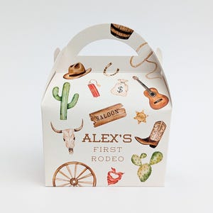 Puede incluir: Una caja de regalo blanca con asa, decorada con ilustraciones de acuarela con temática de vaquero. La caja presenta un sombrero de vaquero, un cactus, una guitarra y el texto "ALEX'S FIRST RODEO".