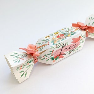 Peut inclure: Un cracker de Noël décoratif avec un motif aquarelle. Le cracker blanc présente une illustration festive, des feuilles vertes, des baies rouges et un ruban rose. Le nom "Lucy" est écrit sur une bannière rose.