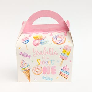 May include: A white party favour box with a pink handle and top, decorated with watercolour illustrations of sweets. The box features the text "Isabella is a Sweet One" in pink and blue, with images of donuts, ice cream, and lollipops.