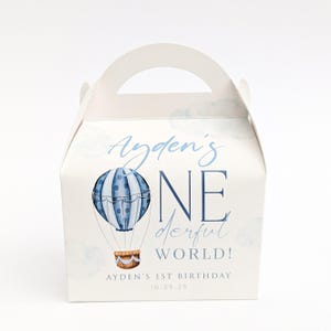 Può includere: Una scatola di cartone bianca con manico, decorata con l'illustrazione di una mongolfiera blu e il testo Ayden's ONEderful WORLD! Ayden's 1st Birthday 10.29.25. Il design include nuvole azzurre e un pallone a righe blu e bianche con cesta.