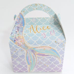 May include: A mermaid-themed gift box with a handle, featuring a mermaid tail design in shades of blue, purple, and gold glitter. The box is decorated with a scale pattern and the text "Alice is 6!"