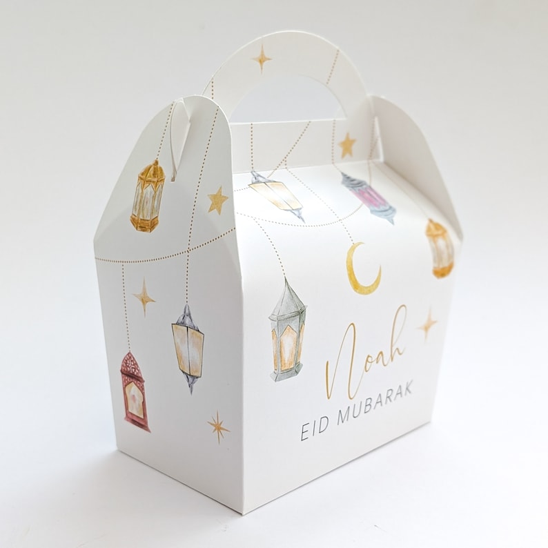 May include: White gift box with a handle, decorated with a crescent moon, stars, and hanging lanterns. The box says "Noah Eid Mubarak".