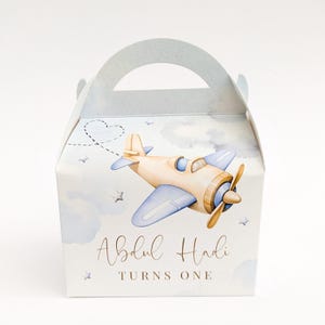 Puede incluir: Una caja de regalo azul claro con asa, con una ilustración de acuarela de un avión vintage. La caja está decorada con nubes, estrellas y el texto "Abdul Hadi TURNS ONE" en escritura dorada.
