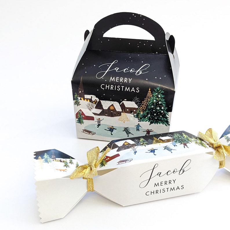 May include: A black and white Christmas gift box and cracker with a winter village scene. The box has a handle and the text "Jacob Merry Christmas". The cracker is tied with a gold ribbon.