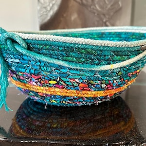 May include: A handmade, oval-shaped bowl with a vibrant, textured design. The bowl features a mix of teal, blue, orange, and yellow fabric strips, with a white rope trim. A teal tassel hangs from the side, adding a decorative touch.