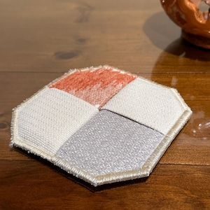 May include: A hexagonal-shaped coaster with a white, gray, and orange fabric design. The coaster has a beige stitched border.