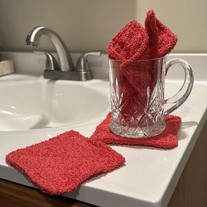 May include: A clear glass mug with a handle, filled with folded red washcloths, sits on a matching red square washcloth. Another red washcloth is next to the mug. The items are on a white countertop near a sink.