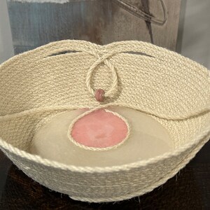 May include: A woven basket made of natural fibers with a pink circle in the center. The basket has a handle made of the same material.