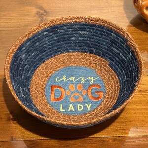 May include: Round woven basket with a blue denim exterior and a tan interior. The center features the words "crazy D&G LADY" in orange and white, with a paw print design.