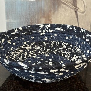 May include: A round, handmade bowl with a navy blue background and a white, speckled pattern. The bowl is made of tightly coiled fabric, creating a textured surface. The bowl is likely used for storage or decoration.