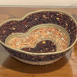 May include: A heart-shaped decorative bowl with a celestial pattern. The bowl is made of coiled rope and fabric, featuring a dark brown background with gold stars and a lighter orange and brown center. The rim is trimmed with a light beige rope.