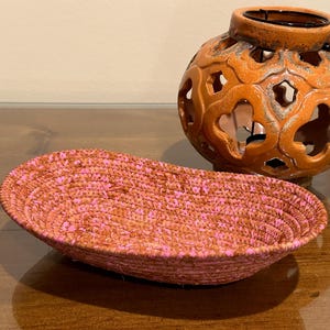 May include: A woven oval basket in shades of pink and orange sits next to an orange ceramic candle holder with decorative cutouts. The candle holder has a dark metal handle. The objects are on a wooden surface.