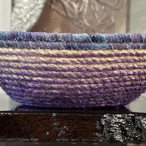 May include: A handcrafted, round bowl made of woven rope. The bowl features a gradient of colors, transitioning from a deep purple at the base to a lighter tan in the middle, and a blue-purple rim. The bowl is sitting on a dark, glossy surface.