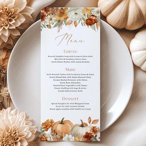 May include: A fall-themed menu card with floral and pumpkin accents. The menu features a list of dishes, including soup, turkey, ham, and dessert options. The card is surrounded by pumpkins, flowers, and a white plate.