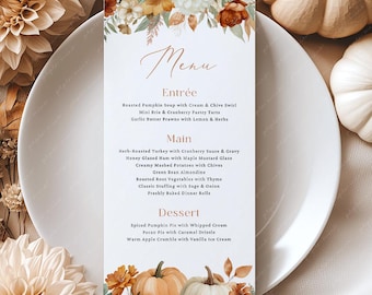 Könnte beinhalten: Eine herbstliche Menükarte mit floralen und Kürbis-Akzenten. Das Menü bietet eine Liste von Gerichten, darunter Suppe, Truthahn, Schinken und Dessert-Optionen. Die Karte ist von Kürbissen, Blumen und einem weißen Teller umgeben.