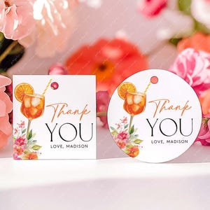Könnte beinhalten: Quadratische und runde weiße Anhänger mit einer Aquarellillustration eines Cocktails mit Orangenscheibe und Strohhalm. Die Anhänger tragen die Aufschrift "Thank You" und "Love, Madison" mit floralen Akzenten. Im Hintergrund rosa und orangefarbene Blumen.