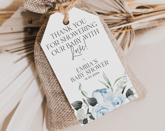 Op de afbeelding: Een klein jute zakje met een wit label eraan bevestigd. Het label heeft blauwe aquarelbloemen en de tekst "Bedankt voor het verwennen van onze baby met liefde! Emilia's Baby Shower 12.05.2024"