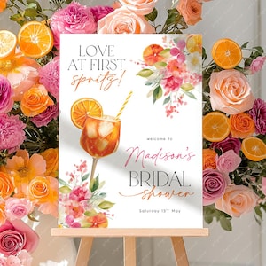 Könnte beinhalten: Ein Schild auf einer Staffelei mit dem Text "Love at First Spritz!" und "Madison's Bridal Shower". Das Schild zeigt eine Cocktail-Illustration mit einer Orangenscheibe. Der Hintergrund ist mit rosa, orangefarbenen und gelben Blumen und Orangenscheiben geschmückt.