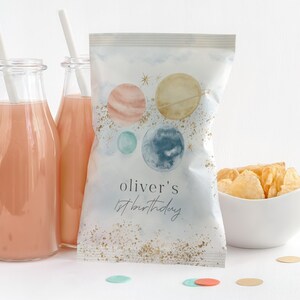 Puede incluir: Una bolsa blanca con un fondo azul acuarela y purpurina dorada. La bolsa tiene un diseño de planetas y estrellas. El texto en la bolsa dice "Oliver's 1st birthday".