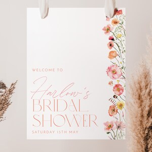 Pu&ograve; includere: Un cartello bianco con testo rosa che dice "Benvenuti al Bridal Shower di Harlow Sabato 15 maggio". Il cartello ha un bordo floreale con fiori rosa e arancioni.