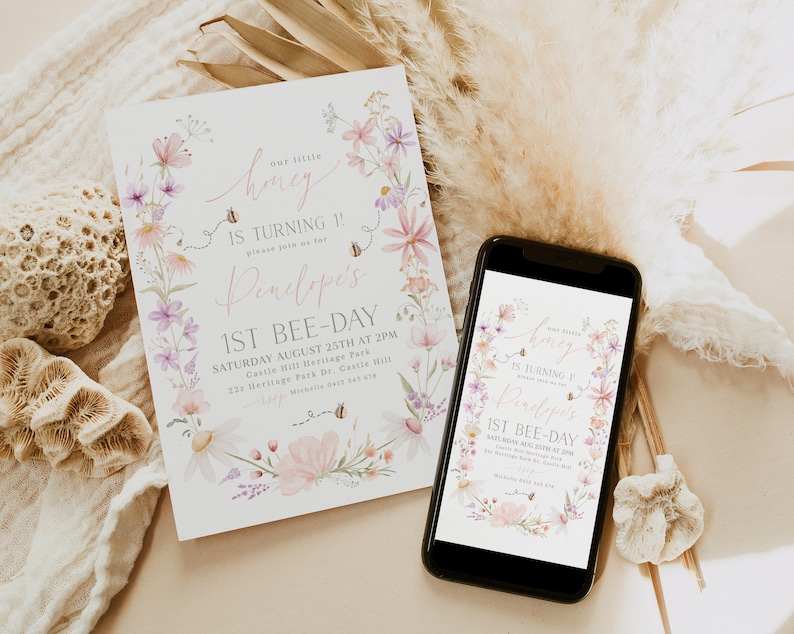 K&ouml;nnte beinhalten: Eine Einladung im floralen Design f&uuml;r eine erste Geburtstagsfeier. Die Einladung zeigt einen Kranz aus rosa und lila Blumen mit Bienen und dem Text "our little honey is turning 1! please join us for Penelope's 1st bee-day" sowie die Partydetails.