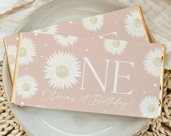 May include: Two chocolate bar wrappers with a pink background, white daisy pattern, and gold foil edges. The wrappers read "ONE" and "Olivia's 1st Birthday!" in white script. The bars are displayed on a light gray plate.