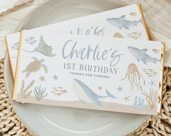 May include: A blue and white chocolate bar wrapper with a watercolor illustration of sea creatures, including a whale, a shark, a turtle, a jellyfish, and a stingray. The text on the wrapper reads "Charlie's 1st Birthday Thanks for coming!"