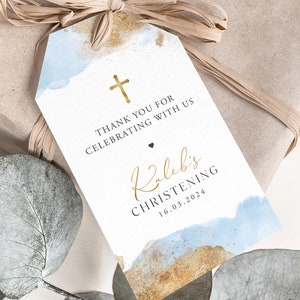 May include: A white gift tag with a watercolor design in blue and gold, featuring the text "THANK YOU FOR CELEBRATING WITH US" and "Kaleb's CHRISTENING 16.03.2024". A gold cross and a small black heart are also present.