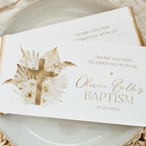 May include: Two white chocolate bar wrappers with gold accents and text. The wrappers feature a gold cross, floral design, and the words "Olivia Belle's BAPTISM" and "THANK YOU FOR CELEBRATING WITH US". The date 30.06.2024 is also printed.