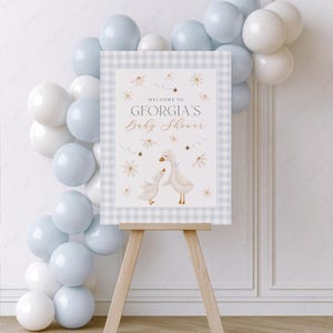 K&ouml;nnte beinhalten: Ein Willkommens-Schild f&uuml;r eine Babyparty mit einem blau-wei&szlig; karierten Hintergrund. Das Schild zeigt zwei wei&szlig;e G&auml;nse mit braunen Augen und Schn&auml;beln und den Text "Willkommen zur Babyparty von Georgia".