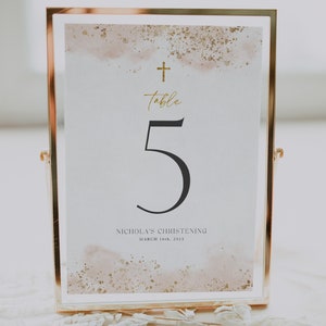 May include: A gold framed table number card with a white background and a gold cross at the top. The number 5 is printed in black and the text "Table" is written in gold script. The text "Nicholas's Christening" and "March 16th, 2023" is printed in black at the bottom.