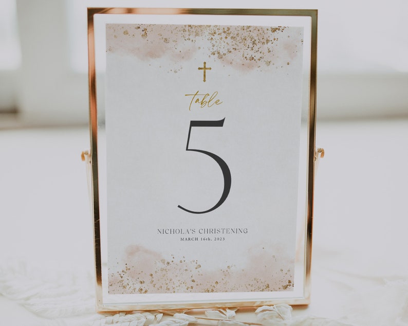 May include: A gold framed table number card with a white background and a gold cross at the top. The number 5 is printed in black and the text "Table" is written in gold script. The text "Nicholas's Christening" and "March 16th, 2023" is printed in black at the bottom.