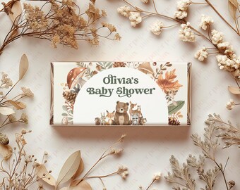 May include: A white chocolate bar wrapper with a woodland theme. The wrapper features a circle with the text "Olivia's Baby Shower" and illustrations of woodland animals, including a bear, deer, and fox. The background of the wrapper is a brown and green pattern of leaves and branches.
