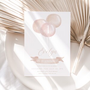 May include: A white invitation card with a watercolor design. The card features three pink balloons with the number "1" on one. The text reads "Evelyn is turning 1" and includes event details. The design is soft and elegant.
